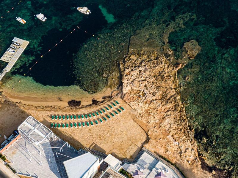 Luftaufnahme eines kleinen Sandstrands mit Liegestühlen neben felsiger Küste und klar blauem Wasser.
