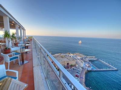 Balkon mit Stühlen und Tischen mit Blick auf einen Hafen und das offene Meer unter klarem Himmel.