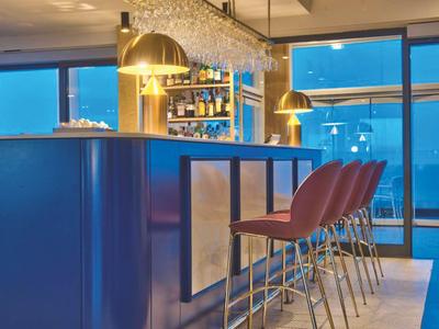 Moderne Hotelbar mit blauer Theke, goldenen Hockern und großen Fenstern mit Blick auf die Stadt.
