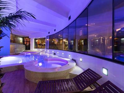 Innenbereich eines Wellnesshotels mit beleuchtetem Whirlpool und Liegestühlen vor einer großen Glasfront.