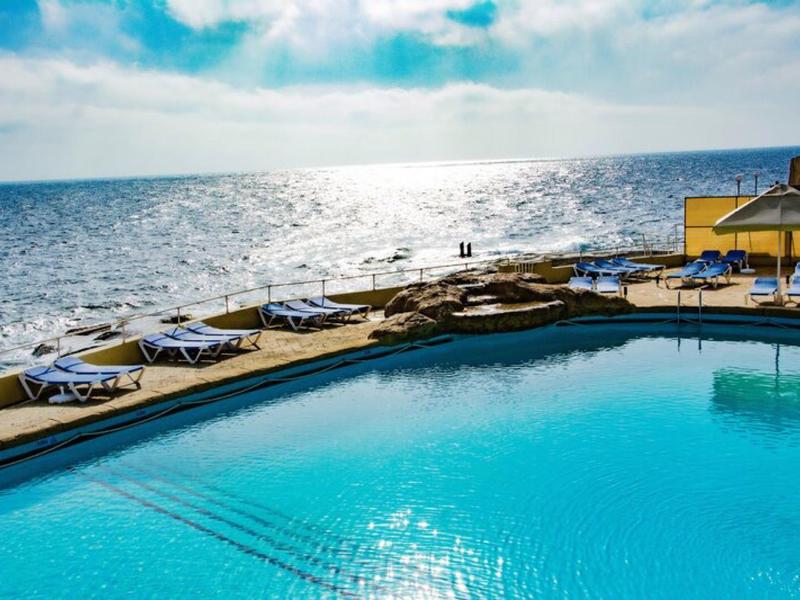 Ein Pool mit Liegestühlen und Blick auf das Meer unter bewölktem Himmel.