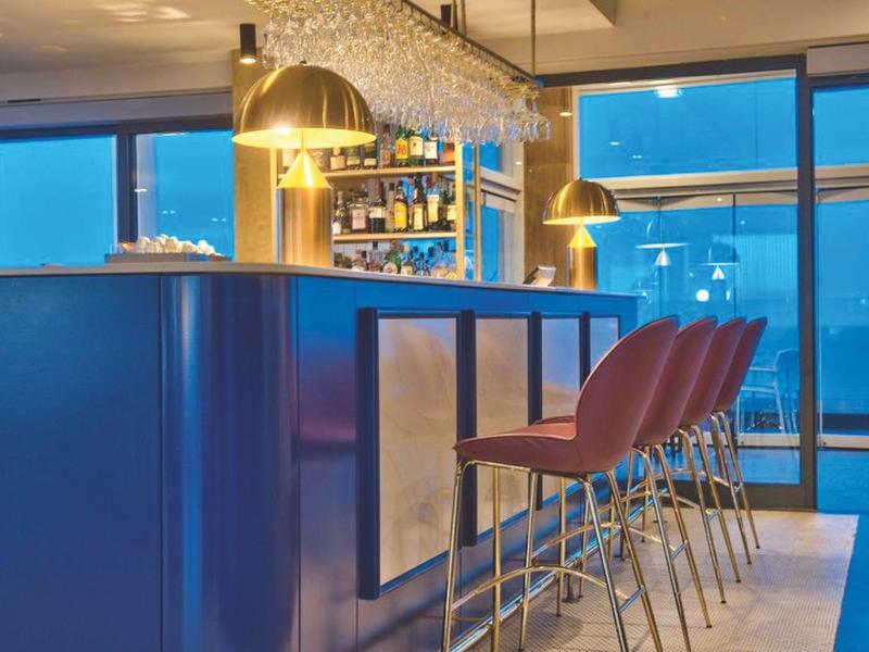Moderner Hotelbarbereich mit blauer Theke, roten Barhockern und großem Fenster mit Blick auf die Stadt.