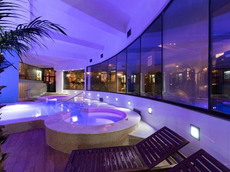 Innenbereich eines Wellnesshotels mit beleuchtetem Whirlpool und Liegestühlen vor einer großen Glasfront.