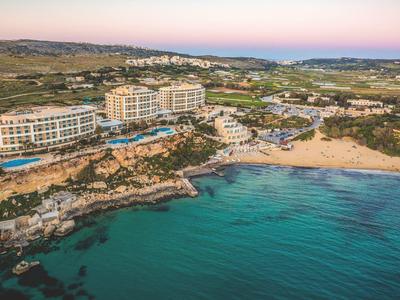 Hotel de lujo en una costa rocosa con playa de arena y mar azul tranquilo al atardecer.