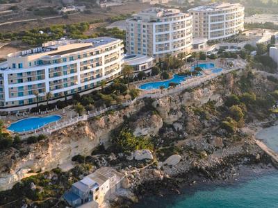 Vista aérea de un hotel moderno frente al mar con piscinas y costa rocosa bajo luz suave.