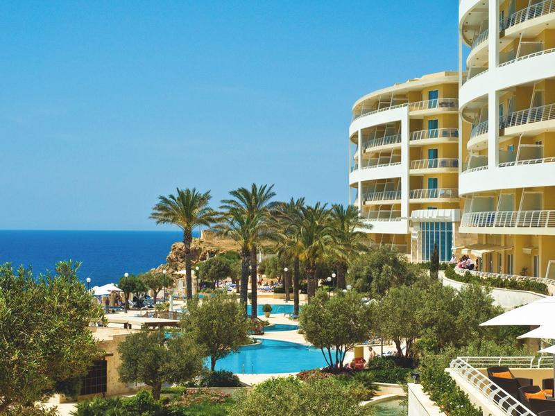 Hotel con balcones redondos, piscina y palmeras junto al mar bajo cielo azul