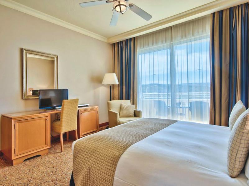 Habitación de hotel luminosa con cama grande, escritorio, sillón y ventana de suelo a techo con cortinas.