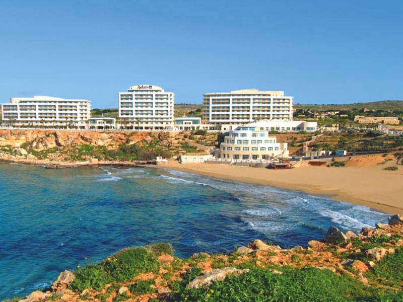 Complejo hotelero con varios edificios junto a una bahía de arena y agua azul clara bajo un cielo despejado.