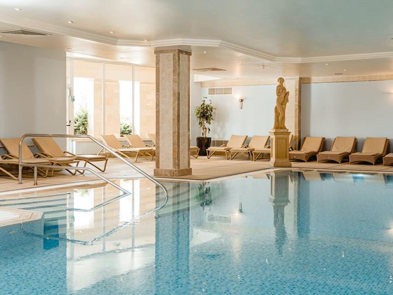 Piscina cubierta luminosa con tumbonas, vistas a ventanas y una escultura en un hotel.