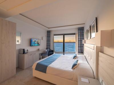 Modernes Hotelzimmer mit großem Bett, Schreibtisch und Balkon mit Meerblick bei Sonnenuntergang.