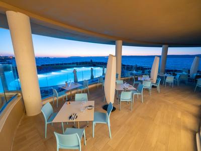 Überdachte Terrasse mit Tischen und Stühlen, Blick auf einen Pool und Meer bei Sonnenuntergang.