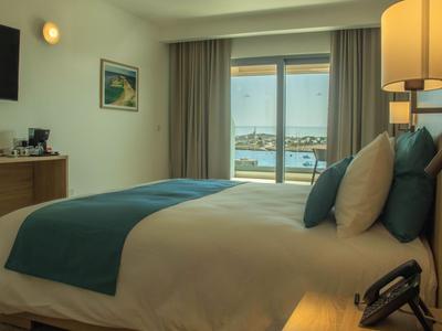 Modernes Hotelzimmer mit Doppelbett, seitlicher Lampe und Blick auf das Meer durch die Balkontür.