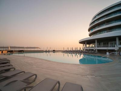 Moderner Hotelpool mit Liegestühlen und Blick auf den Hafen bei Sonnenuntergang.