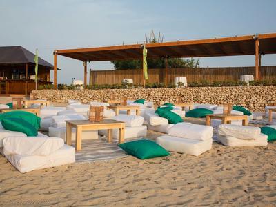 Gemütliche Sitzgelegenheiten mit weißen Kissen und grünen Polstern auf Sand vor einer Strandbar.