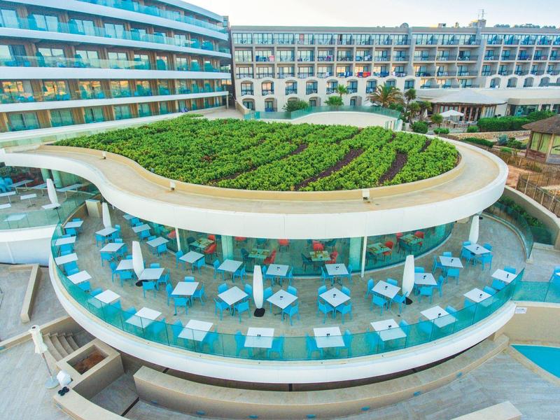 Nowoczesny kompleks hotelowy z zakrzywionym zielonym dachem nad otwartą strefą restauracyjną z stołami i krzesłami.