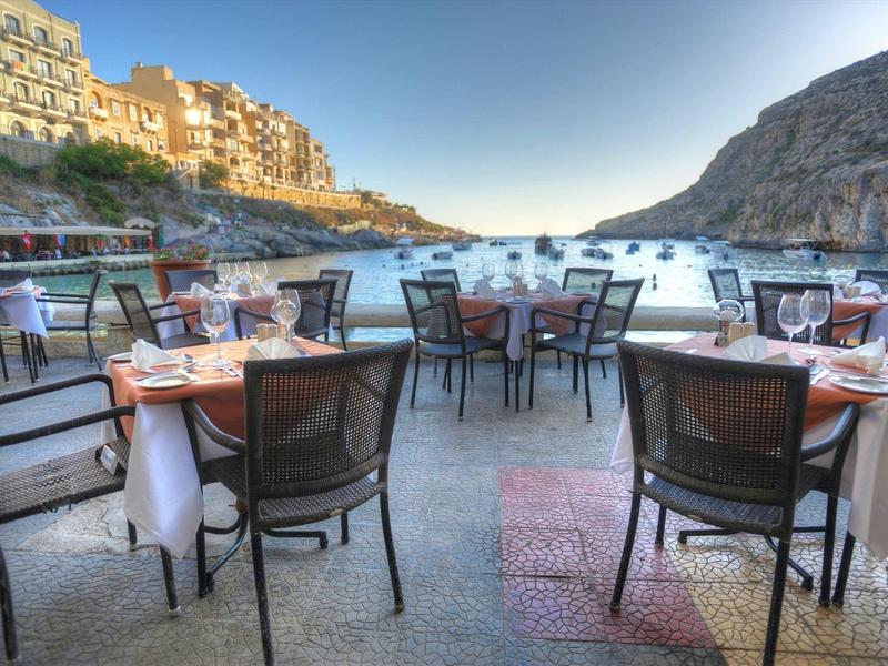 Ristorante all'aperto con tavoli e sedie sul lungomare tra scogliere e edifici.