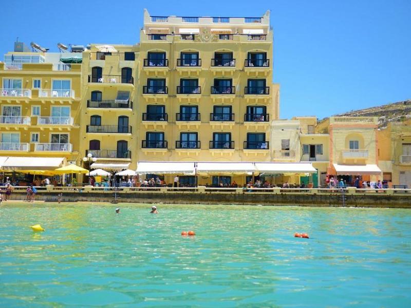 Edificio alberghiero giallo su più piani con balconi vicino all'acqua sotto un cielo blu.