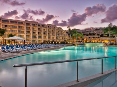 Großer Poolbereich eines Hotels bei Sonnenuntergang mit Liegestühlen und Palmen.