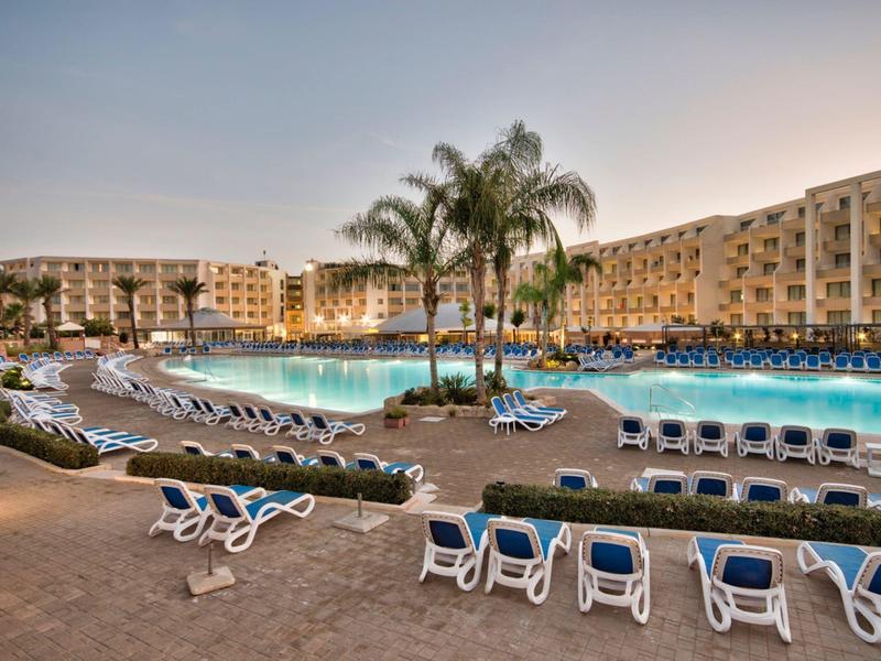 Großer Poolbereich mit Palmen, Sonnenliegen und Hotels im Hintergrund bei Sonnenuntergang.