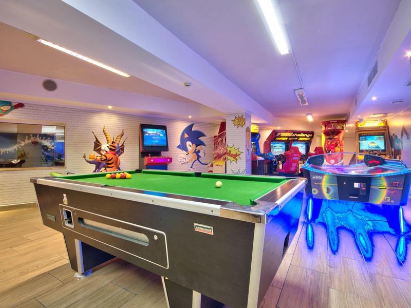 Ein Billardtisch und verschiedene Videospielautomaten in einem farbenfroh beleuchteten Spielzimmer.