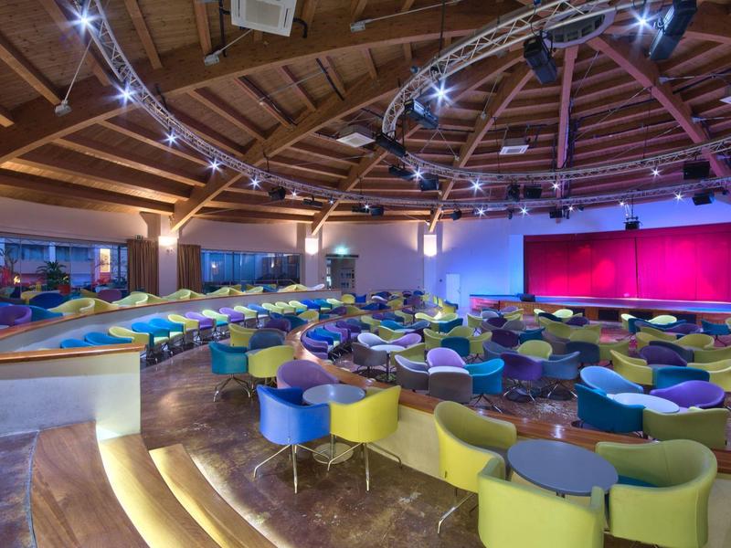 Großer runder Veranstaltungsraum mit bunten Stühlen und Bühne unter Holzdach.