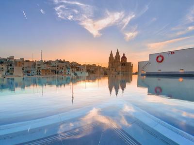 Blick auf eine Stadt mit Gebäuden und Kirche bei Sonnenuntergang, reflektiert in einem ruhigen Wasserbecken.