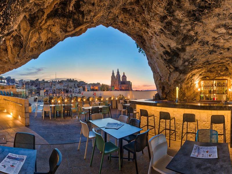 Blick von einer Höhle auf eine beleuchtete Stadt mit Sitzbereich und Bar im Vordergrund.