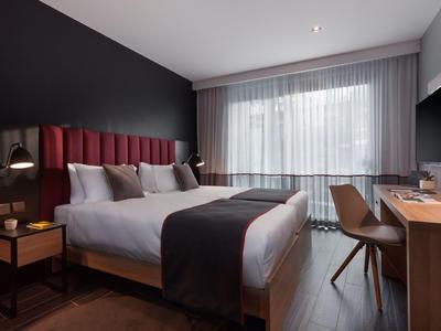 Modernes Hotelzimmer mit großem Bett, Holzschreibtisch und Fenster mit weißen Vorhängen.