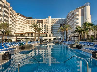 Large hotel with outdoor pool and sun loungers under a clear blue sky.
