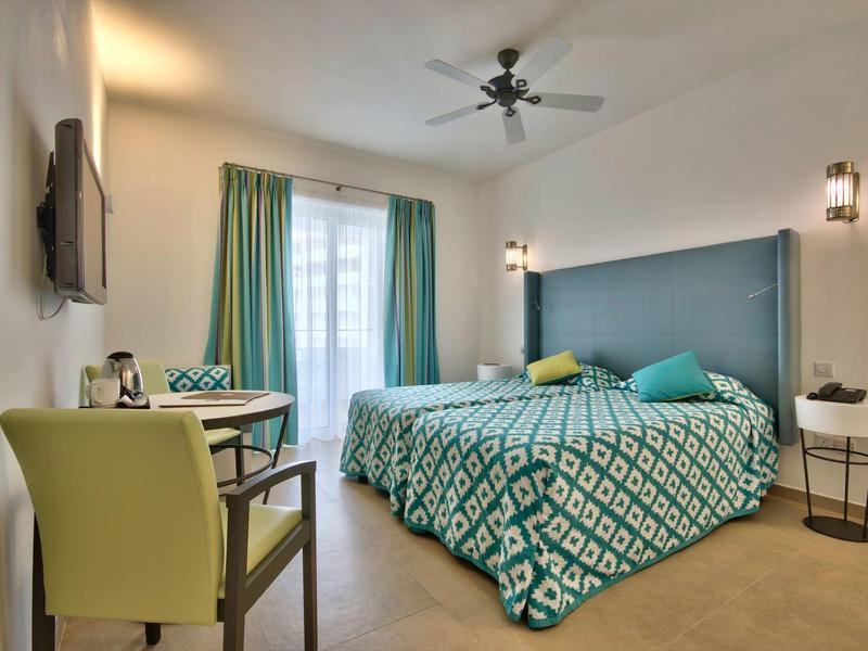 Světý hotelový pokoj s manželskou postelí, zeleným přehozem, křeslem, stolem a balkonem s záclonami.