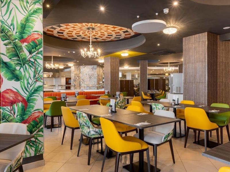 Moderní restaurace s barevnými židlemi a tropickou nástěnnou dekorací.