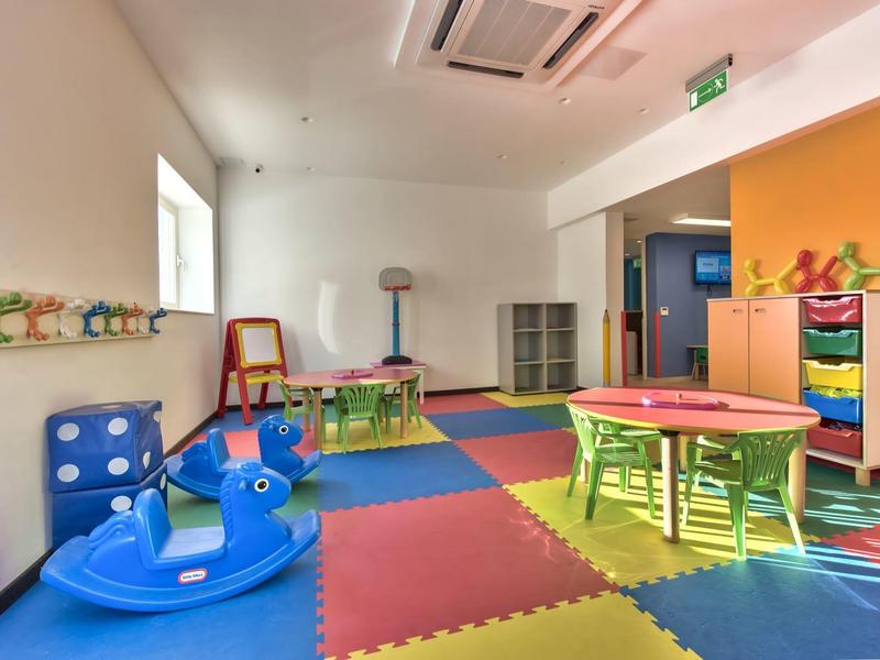 Barevná dětská herna s hračkami, stoly a měkkými podlahovými rohožemi v světlé místnosti.