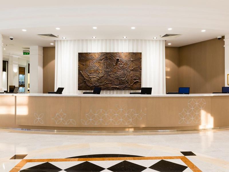 Lobby luminosa de hotel con un largo mostrador de recepción y una pared decorativa al fondo.