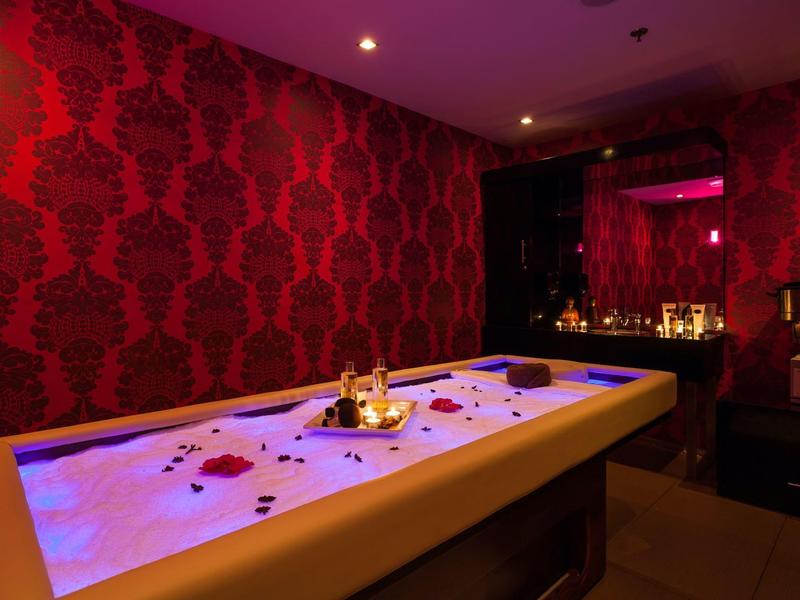 Sala de spa con iluminación ambiental, jacuzzi lleno de pétalos de flores y una vela.