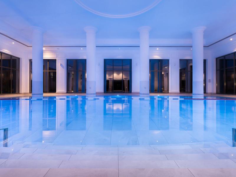 Piscina cubierta con iluminación azul, columnas y grandes ventanas en un hotel.