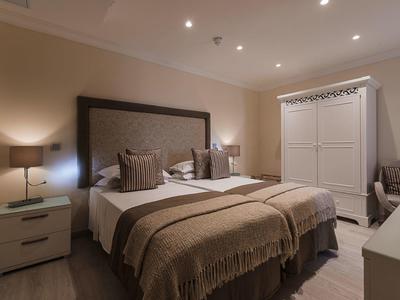 Przytulny pokój hotelowy z łóżkiem podwójnym, stolikami nocnymi, szafą i krzesłem w ciepłych brązowych odcieniach.