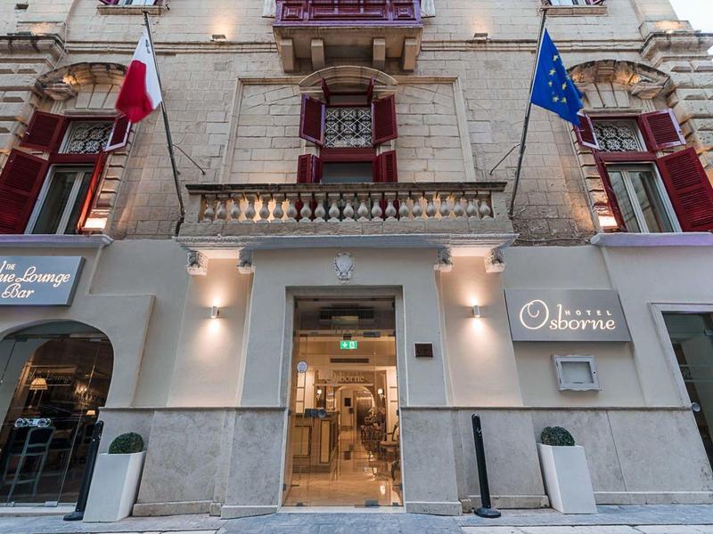 Wejście do hotelu z dwoma flagami, ciepło oświetlonym holem i czerwonymi okiennicami.