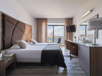 Hotelzimmer mit zwei Betten, TV, Schreibtisch und großer Fenstertür mit Blick auf eine Stadt.