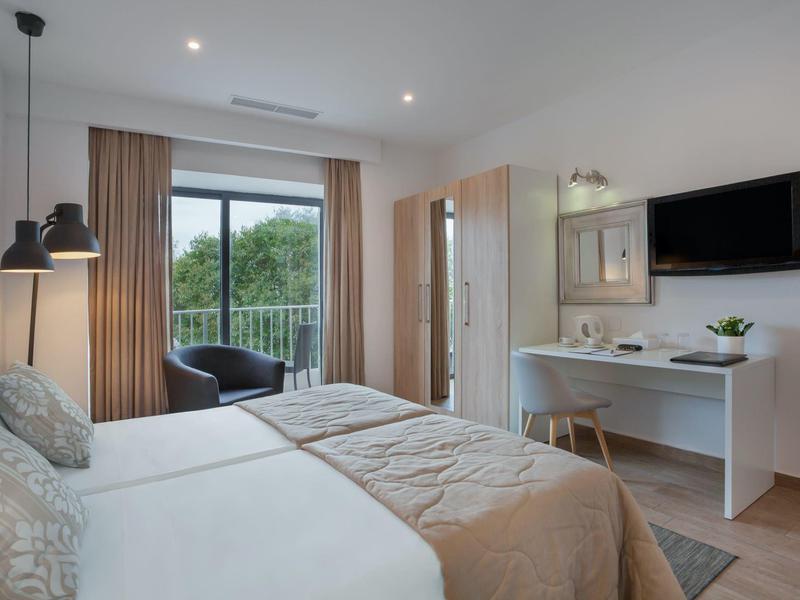Camera d'hotel moderna con due letti, scrivania, TV e balcone con vista sul verde.