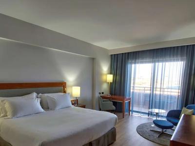 Hell eingerichtetes Hotelzimmer mit Doppelbett, Schreibtisch, Sessel und großem Fenster mit Vorhängen.