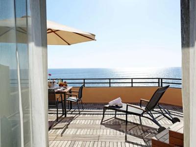 Balkon mit Liegestuhl, Tisch und Stühlen, Blick aufs Meer, sonniger Himmel, helle Holzdielen.