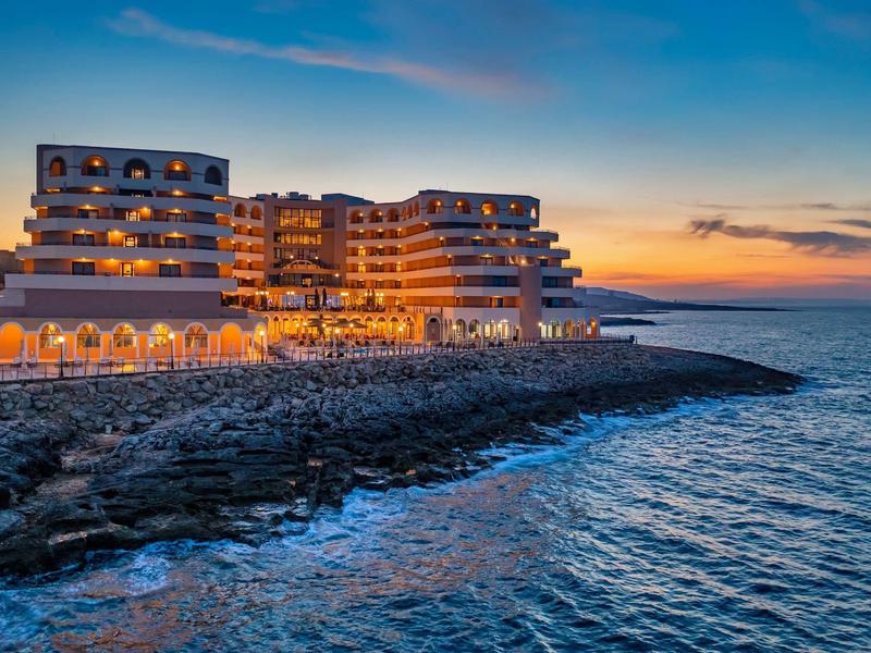 Osvětlený hotel za západu slunce na skalnatém pobřeží s výhledem na klidné moře.