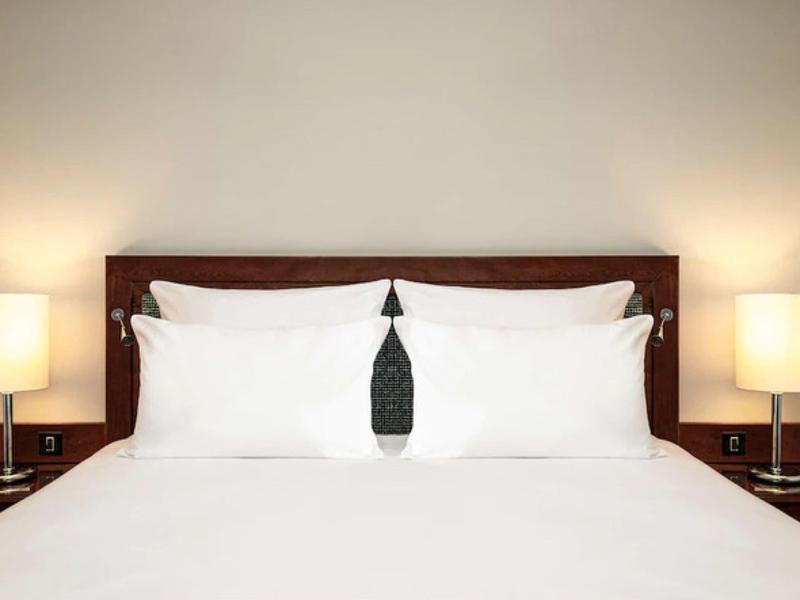 Čistá hotelová postel s bílými polštáři a přikrývkou, dvě zapnuté lampy na nočních stolcích.