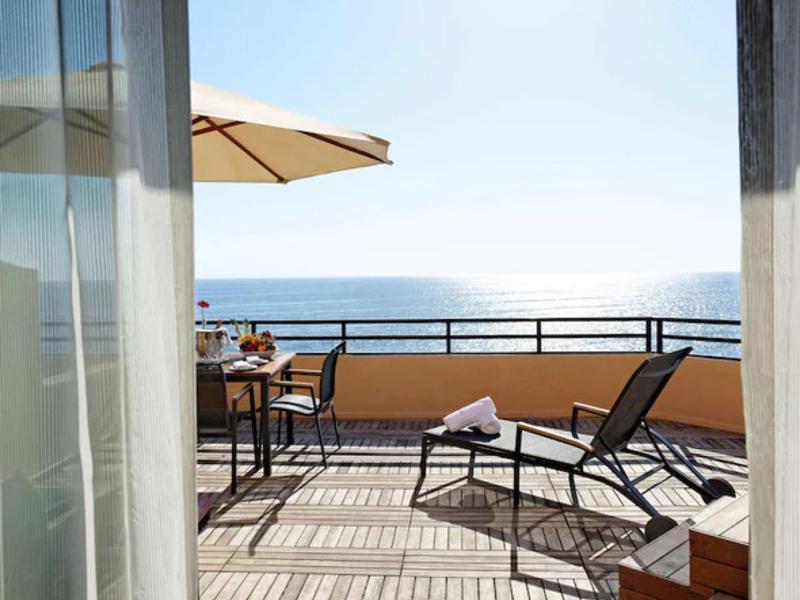 Balkon se stolem, židlemi a lehátkem s výhledem na moře za slunečného dne.