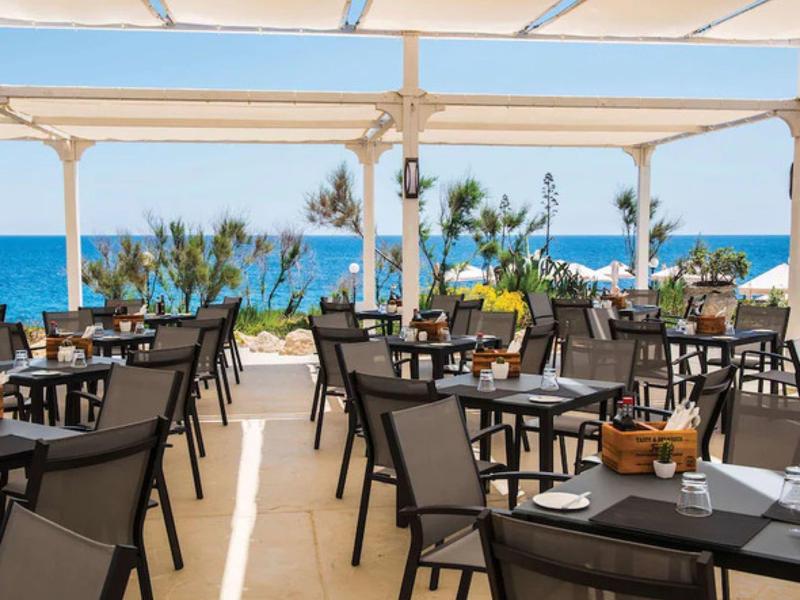 Restaurace na otevřené terase s výhledem na modré moře a rostliny.