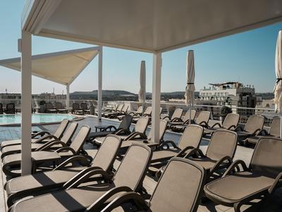 Leere Sonnenliegen unter schattigem Pavillon am Hotelpool bei klarem Himmel.
