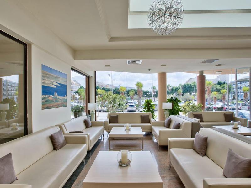 Salon lumineux avec canapés blancs, tables et grande fenêtre donnant sur la ville et la mer.