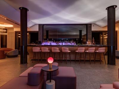 Bar moderna con sgabelli rosa, bancone illuminato e aree salotto accoglienti con luce calda.