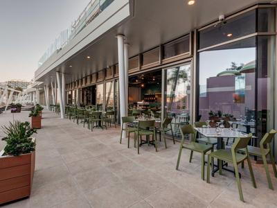 Area esterna moderna di un caffè con tavoli, sedie e vasi lungo una facciata in vetro.