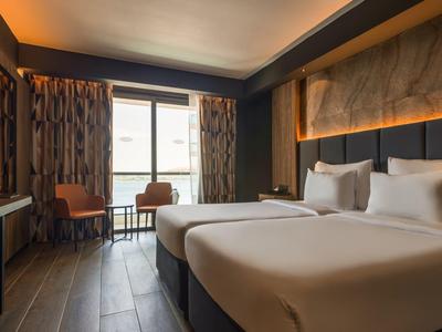 Modernes Hotelzimmer mit zwei Betten, Stuhl, Tisch und Fenster mit Blick nach draußen.